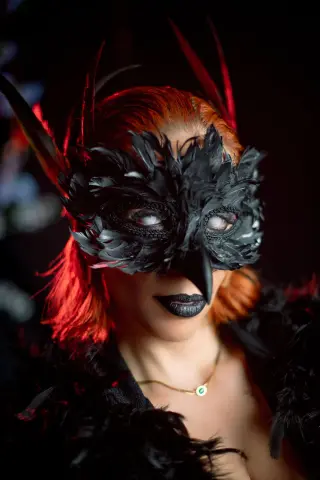 AdelinaVanz wears a black feathered mask and black lipstick with her bright red hair against a dark background.
