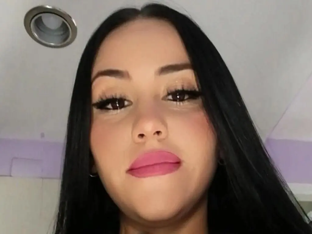 MayaNoiry looking directly at the camera with long, straight black hair and pink lipstick in a close-up headshot.