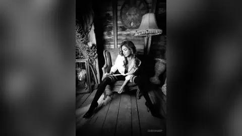 Elegant woman in a white blouse and black leggings sitting in a moody, vintage rustic room.