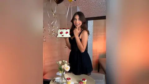 Smiling young woman in a black lace dress holding a birthday cake in a warm, festive indoor setting.