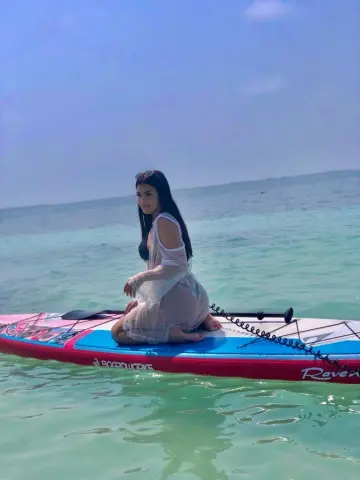 Young woman in a white lace cover-up relaxing on a paddleboard in turquoise ocean water under a clear sky.