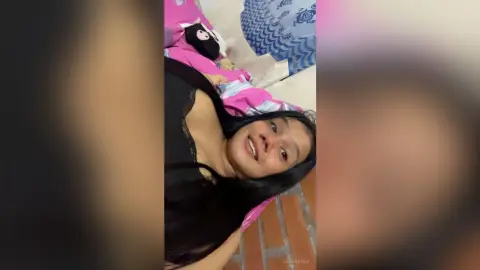 Smiling young woman in black loungewear relaxing on a pink bed with plush toys in a cozy bedroom setting.