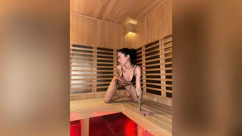 Woman in black bikini posing in a warm, wooden sauna with soft ambient lighting.