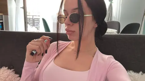 AriaVex wears sunglasses and a pink zip-up top while sitting on a dark grey sofa.