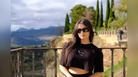 Young woman in black crop top and sunglasses standing on a scenic mountain overlook during a sunny day.