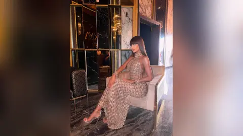 LanaRhode sits on a white chair wearing a sparkly, floor-length halter dress and clear heels in a luxury room.