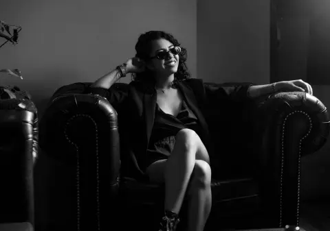 Stylish woman in a black blazer and sunglasses relaxing on a leather sofa in moody black and white lighting.