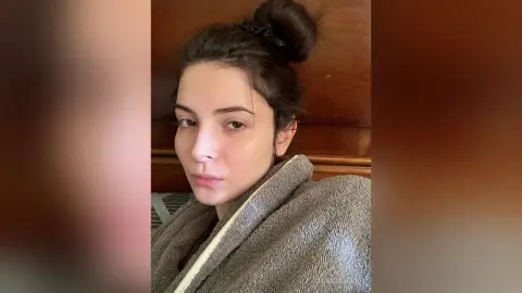 Young woman with hair in a bun wearing a cozy grey bathrobe in a warm indoor setting.