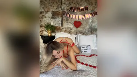 Blonde woman in red lingerie lying on a bed with birthday decorations in a romantic bedroom setting.