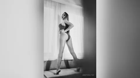 Elegant woman in a lace bodysuit posing by a window in a moody black and white indoor setting.