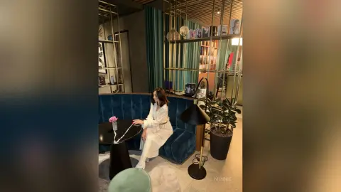Elegant woman in a white outfit sitting on a blue velvet sofa in a chic, modern lounge setting.