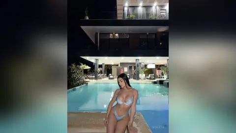 Young woman in a light blue bikini relaxing by a luxury rooftop swimming pool at night.