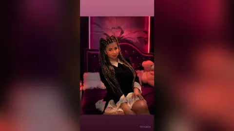 Woman with long braids in a black shirt and white skirt sitting on a bed in moody red lighting.