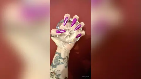 Close-up of a tattooed hand with long purple stiletto nails holding a crystal glass against a red background.