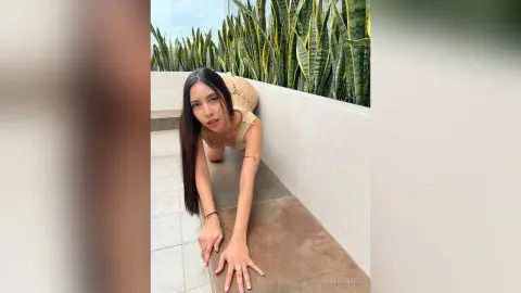 IrinaRussel poses with long dark hair, wearing a tan top while leaning forward on a stone ledge outdoors near green plants.