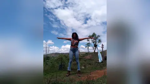 Cheerful woman in a striped tank top and jeans stretching her arms out in a sunny green meadow under a blue sky.