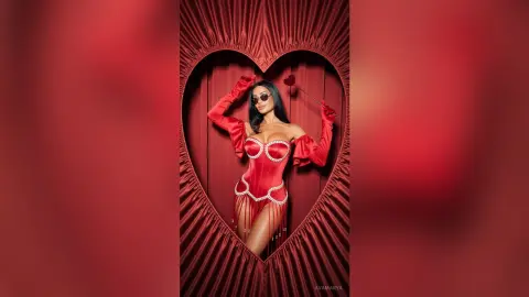 Glamorous woman in a red corset bodysuit posing inside a heart-shaped velvet curtain frame.