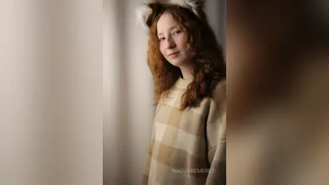 Redheaded woman in a cozy beige checkered sweater smiling in a warm, soft-lit indoor setting.