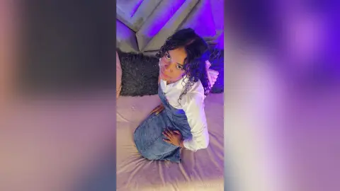 NaomiVelour poses on a bed wearing a white long-sleeve shirt and denim overalls, looking up at the camera with her curly hair down.