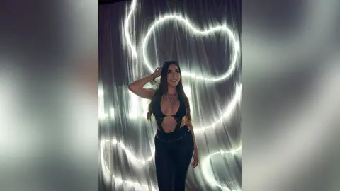 Young woman in a black cutout bodysuit posing against a glowing neon light backdrop in a moody setting.