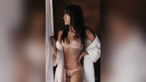 Young woman in white lace lingerie and a bathrobe standing by a window in a moody, rustic room.
