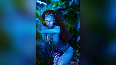 Blue Na'vi woman cosplayer in tribal jewelry posing in a lush, mystical tropical jungle setting.