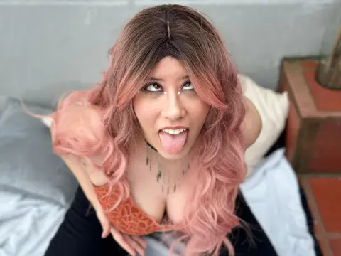 Izzumi sticks out her tongue while wearing an orange lace bra and looking up at the camera with pink wavy hair.