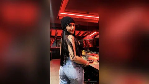 Young woman in a black beanie and denim jeans posing in a moody, red-lit restaurant setting.