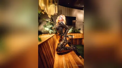 Glamorous blonde woman in a sleek black bodysuit and high boots posing in a moody, modern lounge setting.