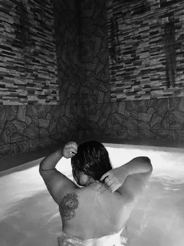 Relaxed woman with a back tattoo relaxing in a steaming indoor hot tub against a textured stone wall.