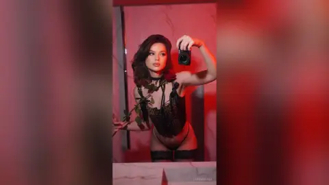 Young woman in black lace lingerie holding a camera in a moody red-lit room.