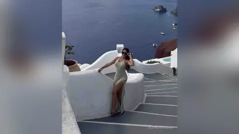 Elegant woman in a long white dress posing on a Santorini balcony overlooking the blue Aegean Sea.