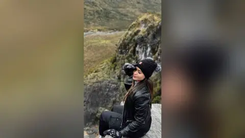 Woman in black leather jacket and beanie enjoying a scenic view in a misty mountain landscape.