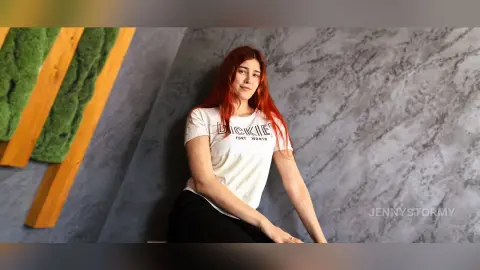 Smiling redhead woman in a white Dickies t-shirt sitting against a textured grey wall in a modern interior.