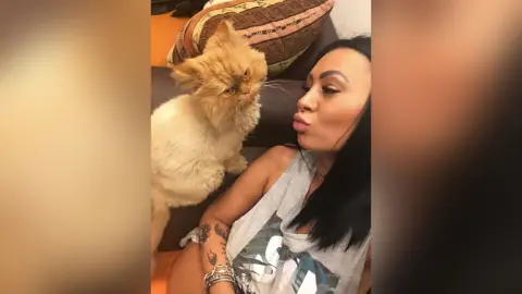 DeeaAnderson pouts her lips while sitting next to a fluffy tan cat in a grey tank top.
