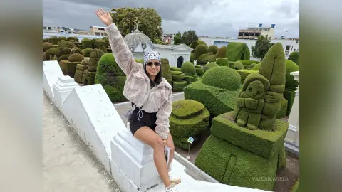 Woman in a fuzzy beige jacket and beanie waving in a lush green topiary garden under a cloudy sky.