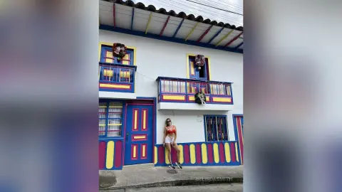Woman in a red bikini and white shorts standing before a colorful colonial building with blue and yellow accents.