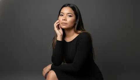 XinemaBonet poses with her hand against her face, wearing a long-sleeved black dress against a dark grey background.