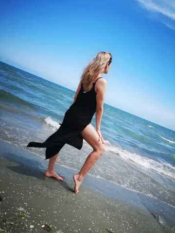 Blonde woman in a black summer dress walking on a sunny beach with ocean waves.