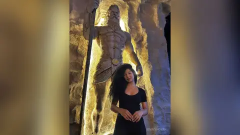 Young woman in a black top standing before a massive, glowing statue of a muscular mythological god in a cave.
