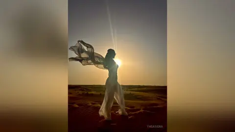 Silhouette of a woman in a flowing white dress walking through desert dunes at sunset.