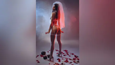 Sensual woman in white lace lingerie and veil posing in a dreamy, smoky studio setting with red rose petals.