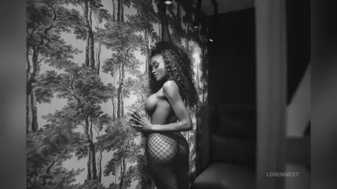 Moody black and white portrait of a woman in fishnet tights posing against vintage forest wallpaper.