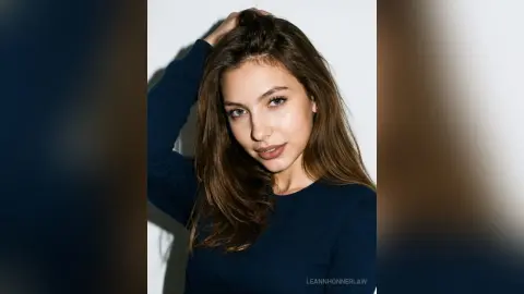 Young woman with long brown hair wearing a navy blue long-sleeve shirt in a bright studio setting.