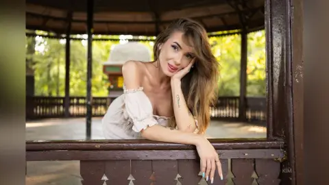 Beautiful woman in a white off-the-shoulder blouse relaxing in a rustic wooden gazebo amidst lush green trees.