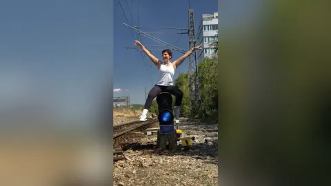 JaneHope — JaneHope sits on a black object on the train tracks with her arms outstretched under a clear blue sky.