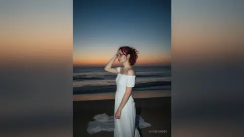 Red-haired woman in a white flowing dress standing on a beach at sunset with a dreamy, serene mood.