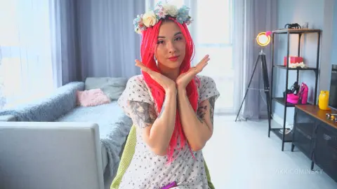 VikkiCominsky poses with her hands near her face, wearing a white patterned top and a light blue floral crown in a bright room.