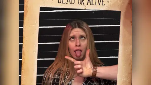 Playful blonde woman in a plaid shirt making a funny face in front of a Dead or Alive chalkboard sign.