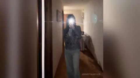 Woman in a dark long-sleeve top and grey trousers taking a moody mirror selfie in a dimly lit hallway.